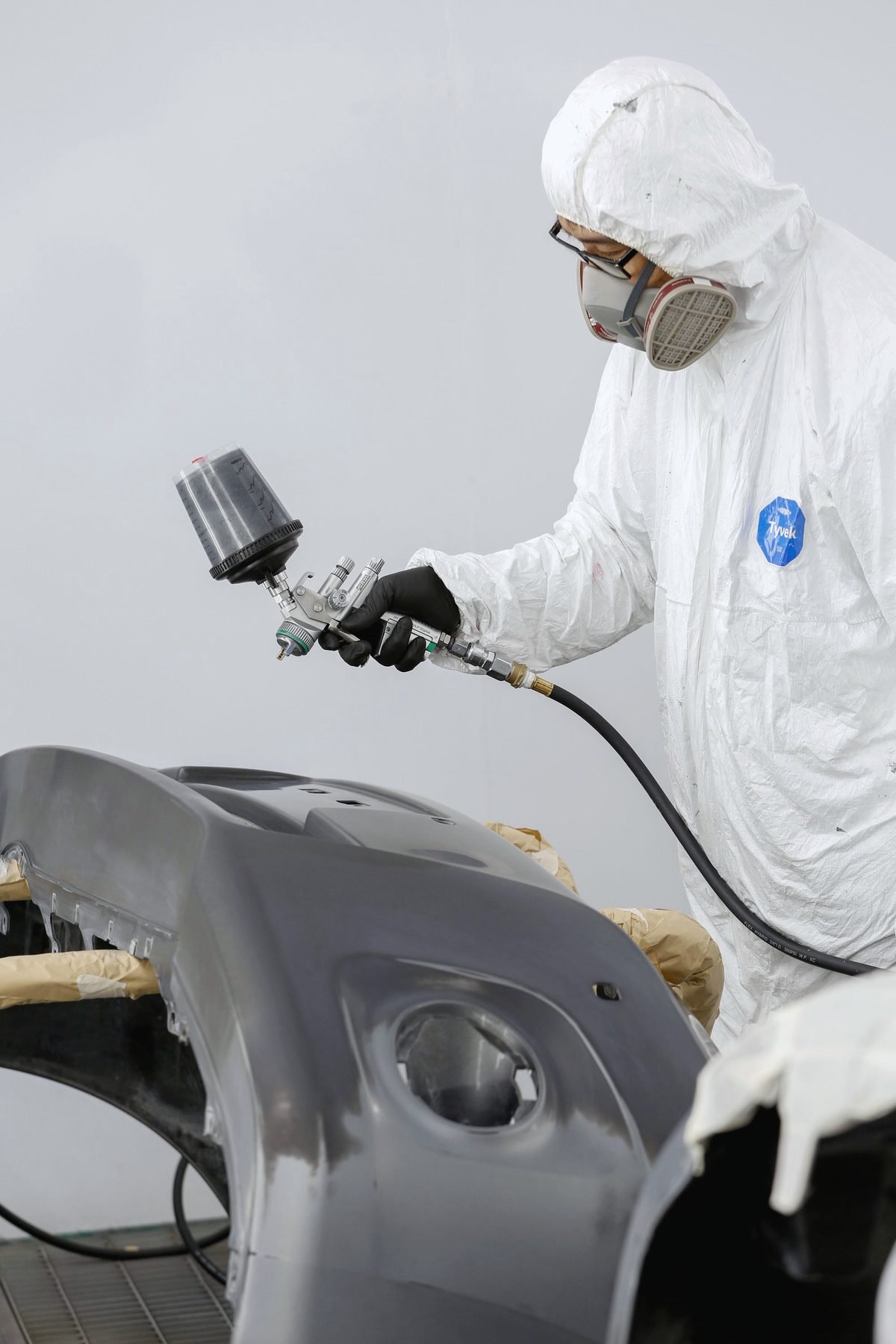 A painter in a white suit and respirator spraying a grey primed bumper
